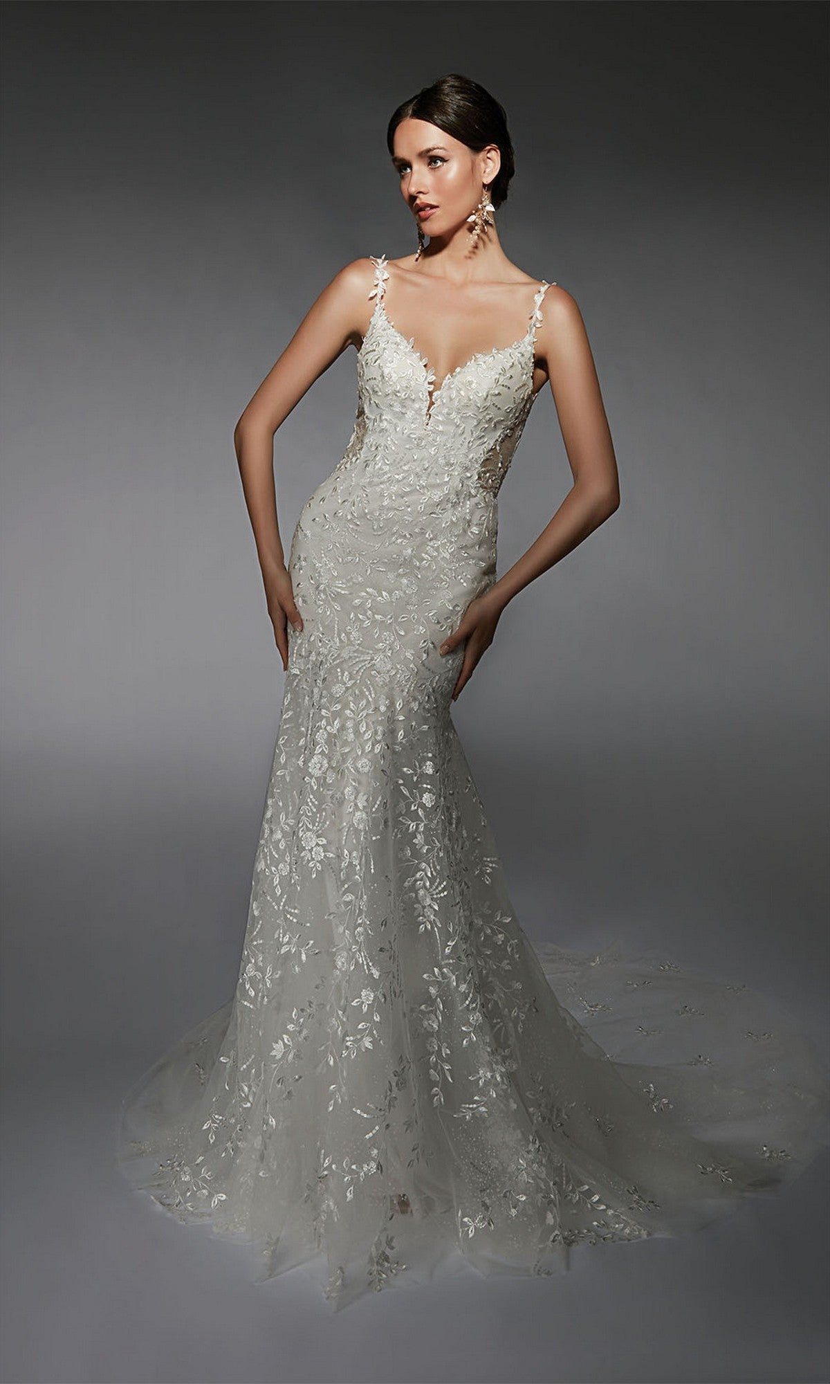 A woman stands against a dark background wearing the Long White Dress: Alyce 7122—a sleeveless wedding gown with a fitted bodice, v-neckline, thin straps, floral lace appliqués, and a long flowing train.